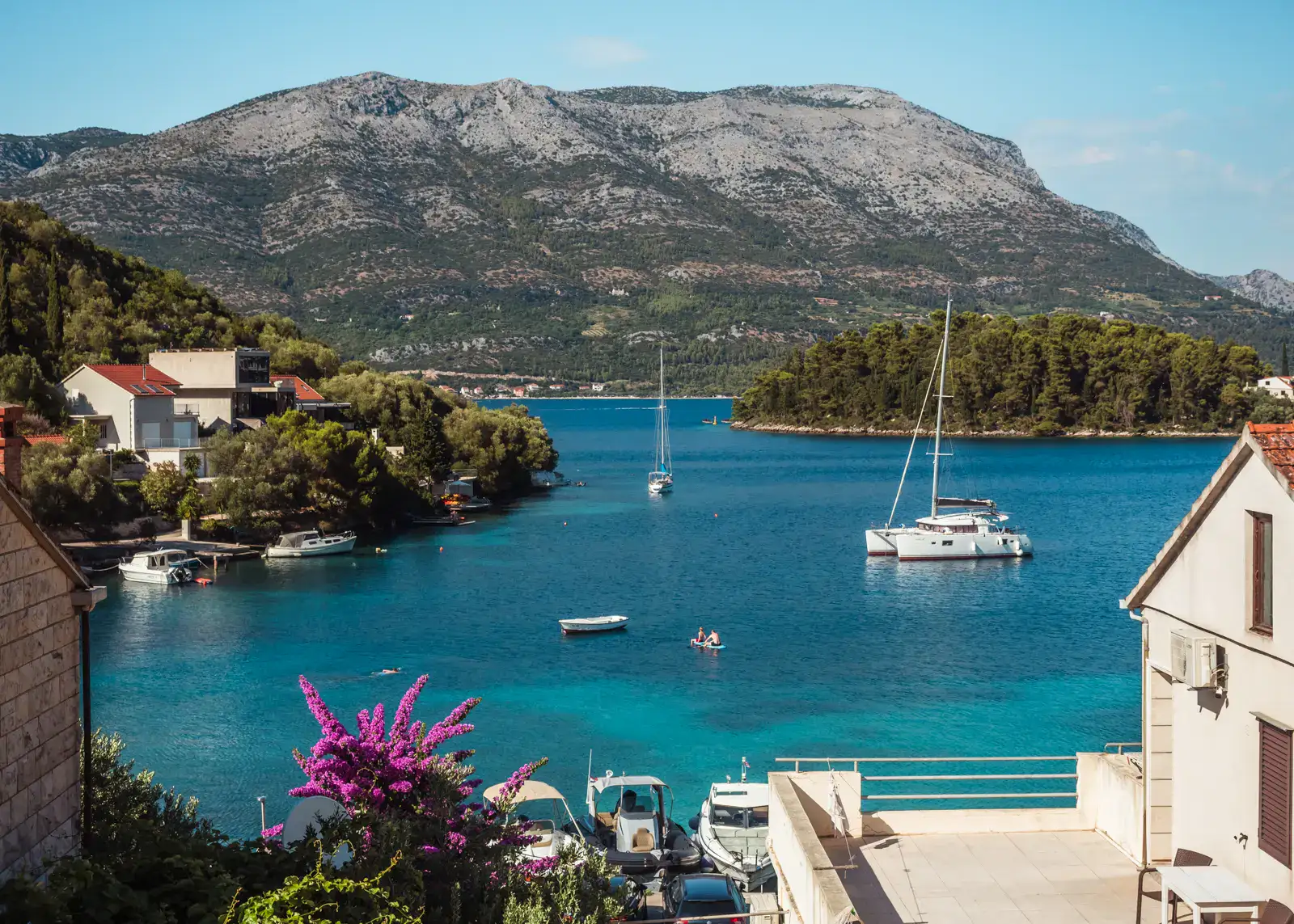 View from our holiday apartment in Korčula, of a mountain in the background, a white catamaran on turquoise water, a little green filet on the right, purple bougainvillea in the foreground, on a sunny day in Korčula, Croatia.