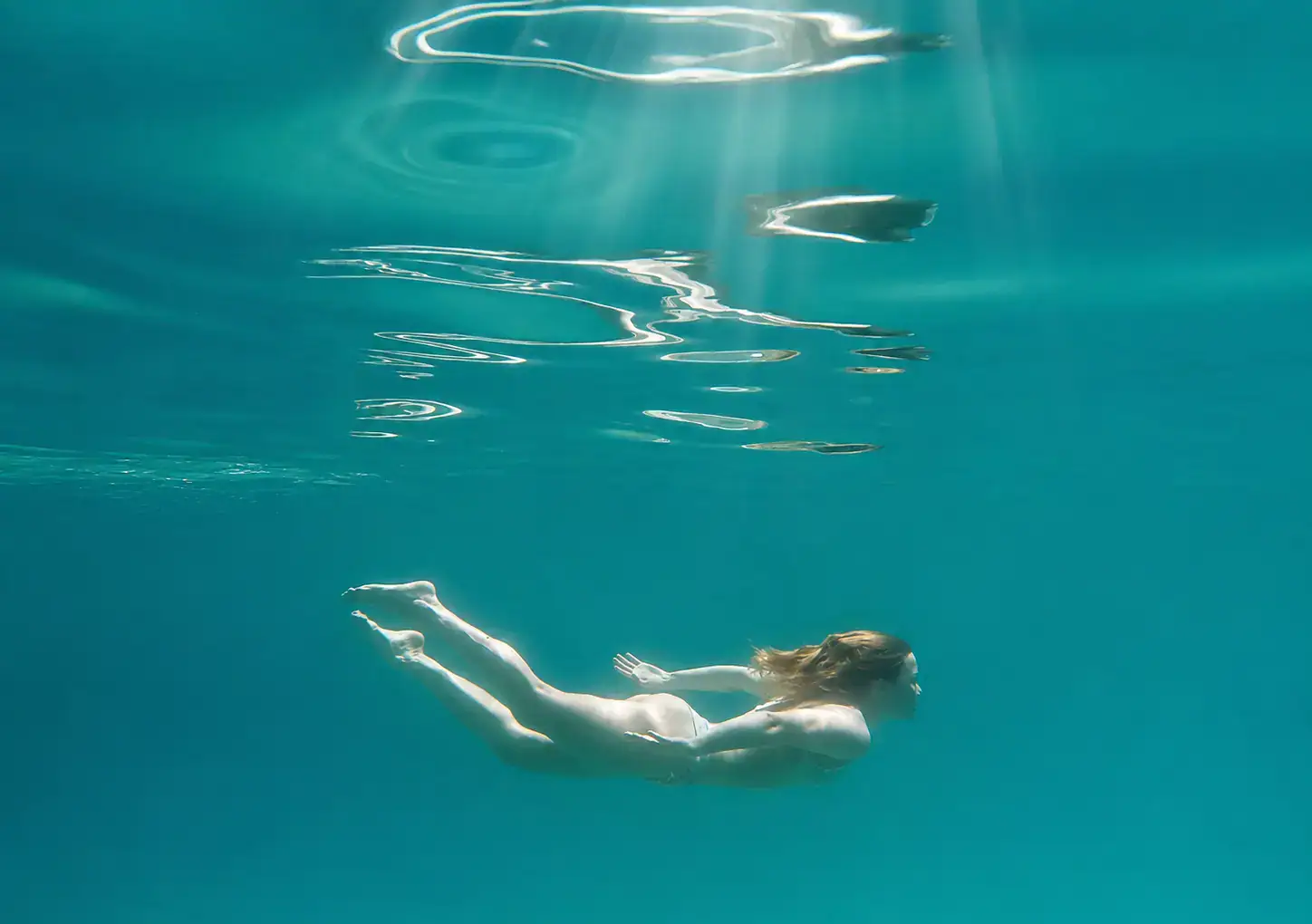 Woman with light skin, red hair, swimming underwater in turquoise/blue ocean, reflection in the water and sun rays shining down, in Žrnovska Banja, Korcula.