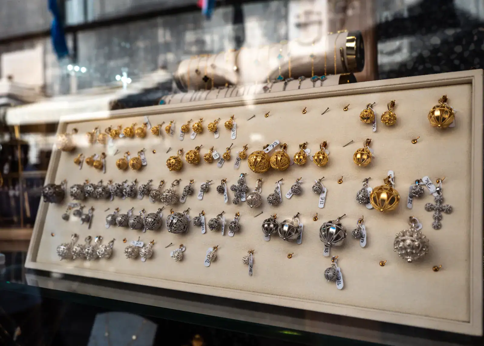 Looking through the window of a jewelry shop on a long display of ornate ball pendants called Botuni in different sizes and gold and silver.