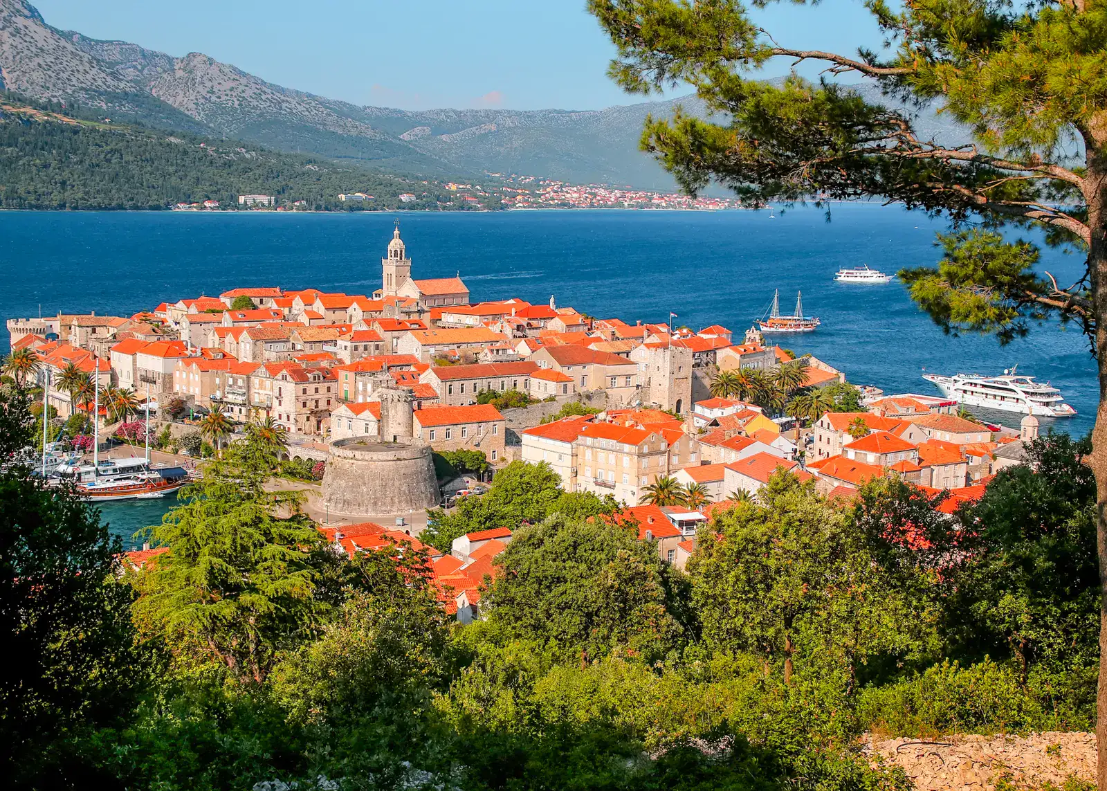 Korcula Old Town seen from above, sandstone houses with orange roofs, framed by green trees, with blue water and a mountain in the background on a sunny day.