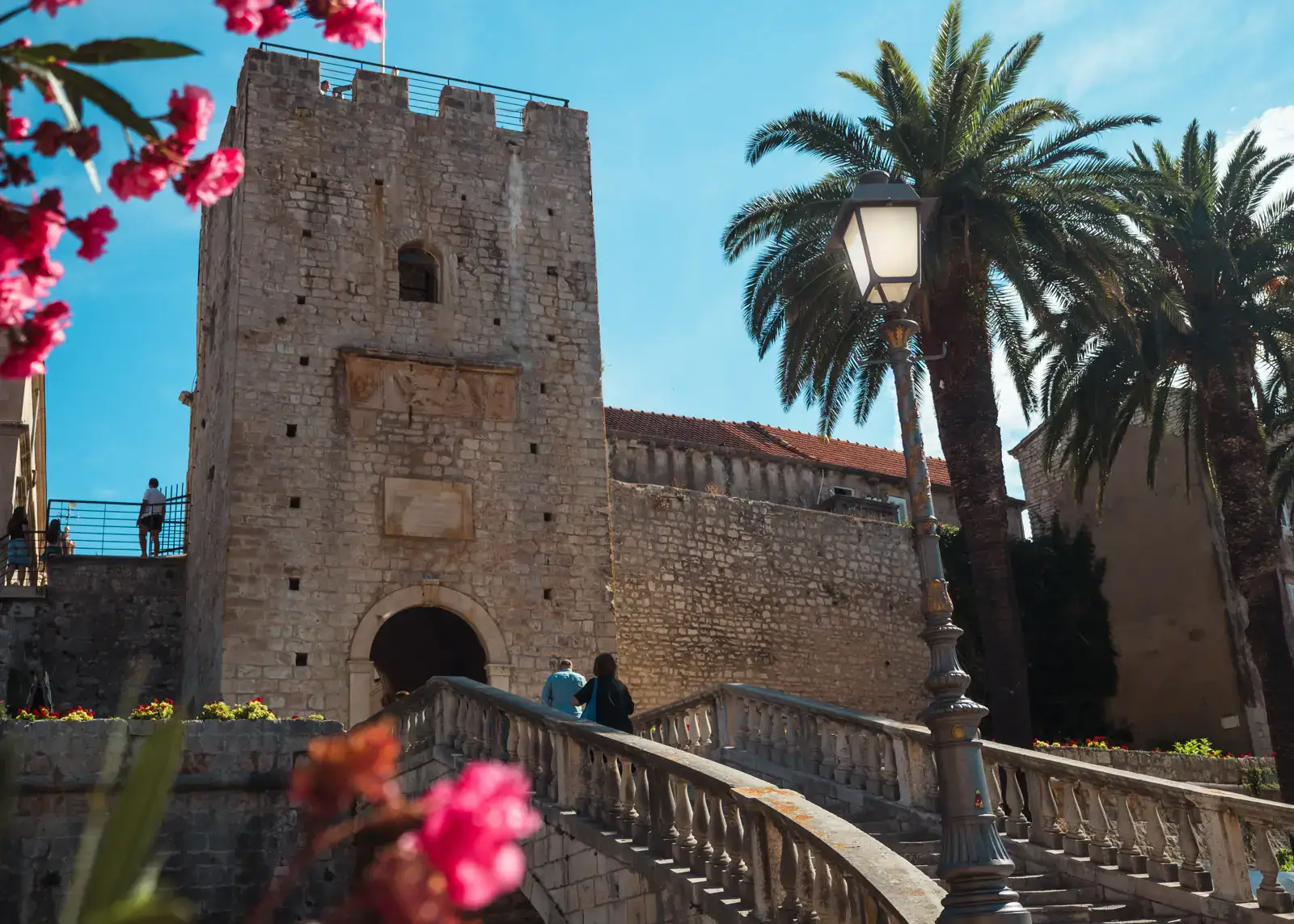 Looking up a stone staircase towards the entrance of Korcula Old Town under a tower, pink flowers in the foreground, blue skies behind. Top things to do in Korcula.