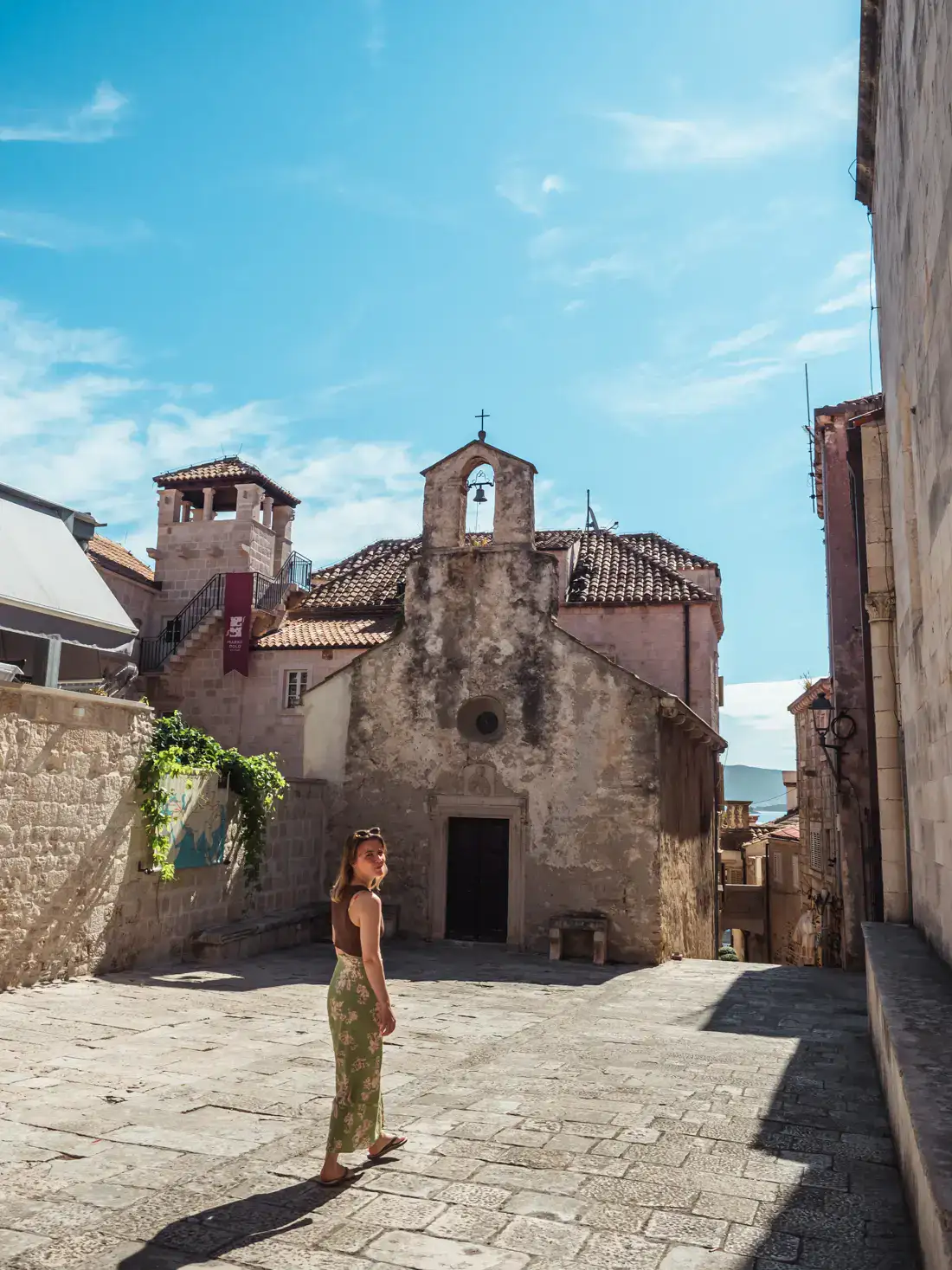 Woman wearing a brown top and long, green skirt, walking in a square with a small church in the background in Korcula Old Town on a sunny day.