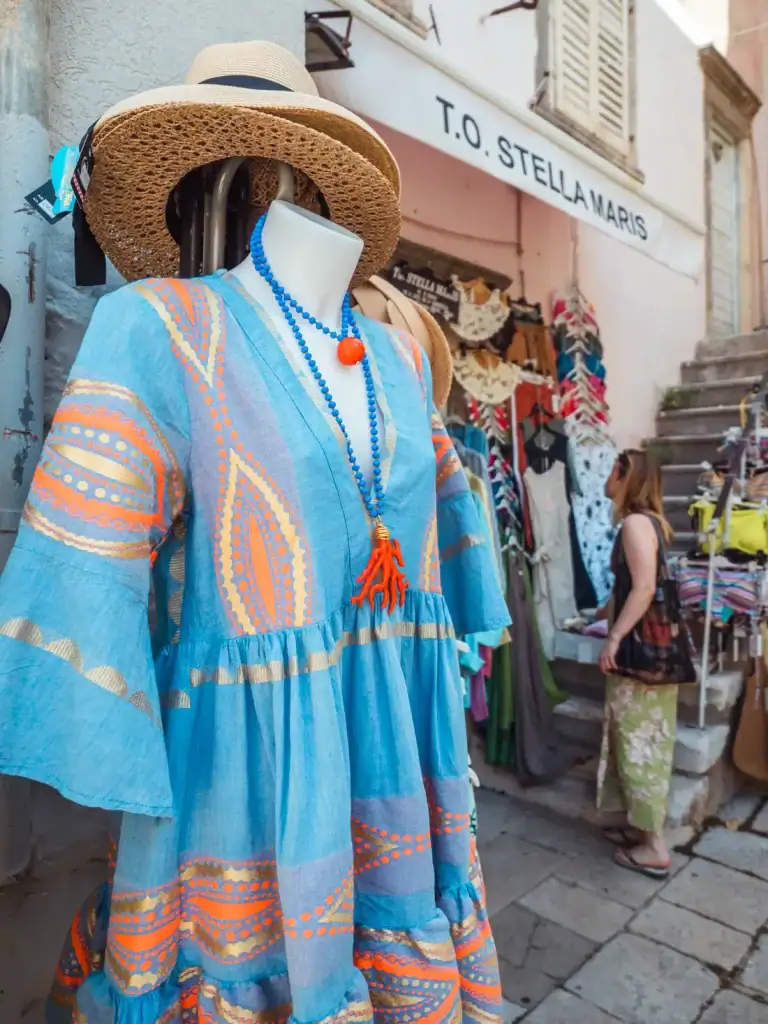 Close up of a blue summer dress with red snd orange pattern on a mannequin with a brown straw hat, woman wearing a brown top and green skirt, looking at clothing by the entrance in the background while shopping in Korcula Town.