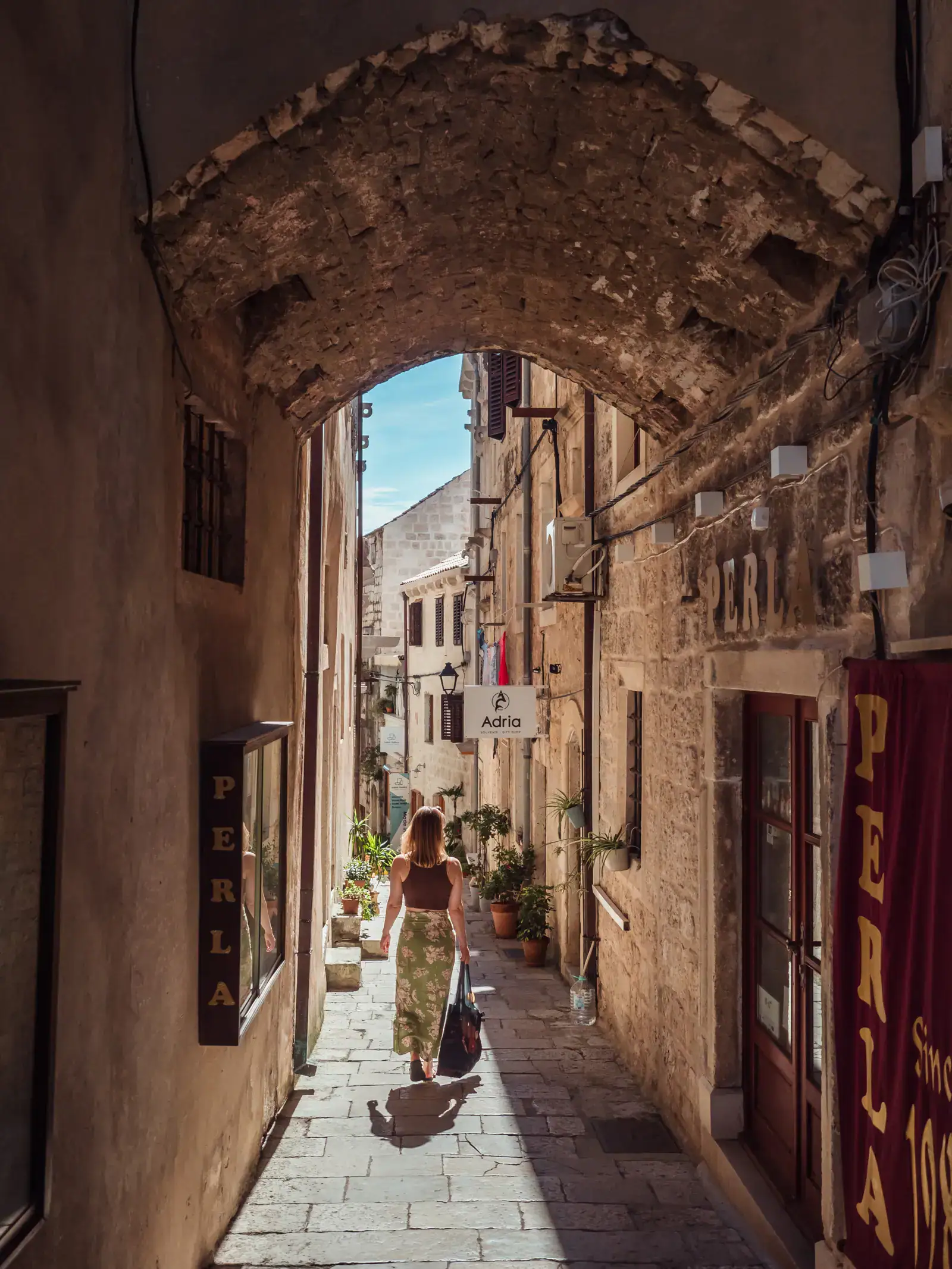 Woman, wearing a brown top and green skirt, holding a black bag, walking down a narrow alley under an arch, with the entrance to a pearl store on the right in Korcula Old Town on a sunny day.