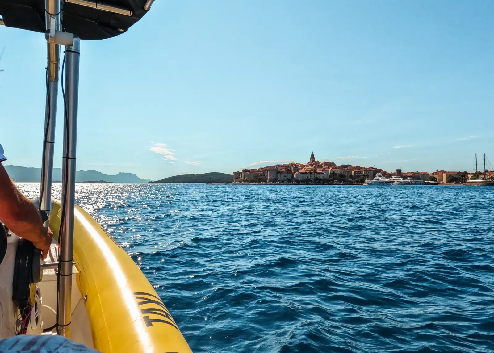 Photo of Korcula Old Town in the distance taken from a yellow taxi boat on a sunny day with blue skies. How to get around Korcula.