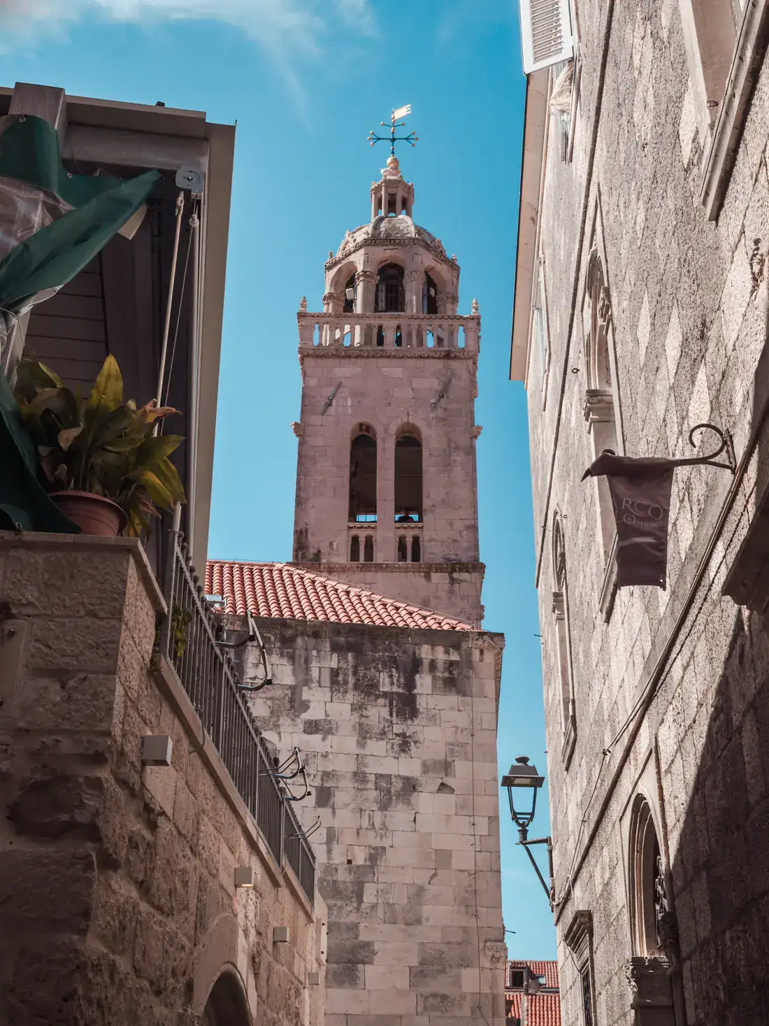 Walking down an alley in Korcula Town, looking uo at the bell tower of St Marks Cathedral against a blue sky.