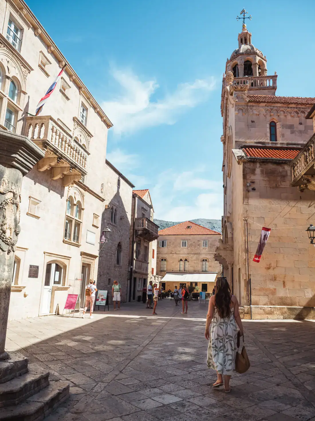 Woman with long, brown hair, wearing a white dress with green pattern, walking in a square towards St Marks Cathedral on a sunny day in Korcula Town.