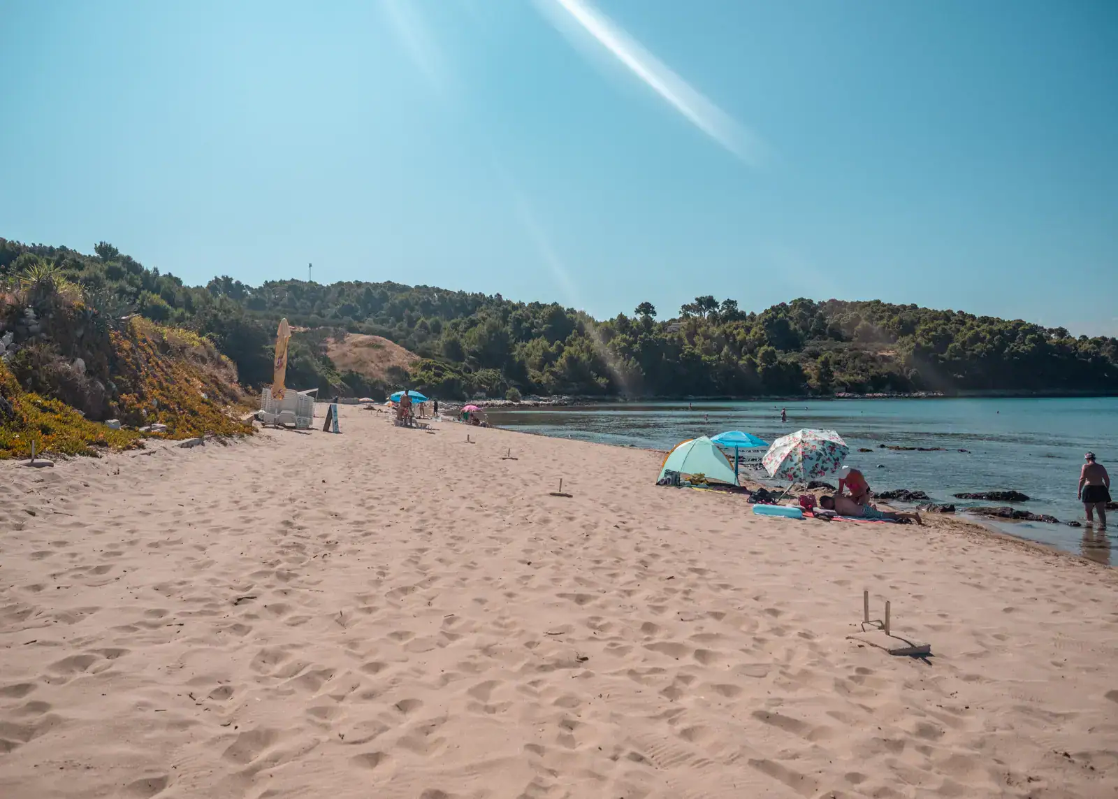 View of Vela Pržina Beach early in the morning on a sunny day with blue skies, a sun ray shining in, beige, soft sand, a few colorful umbrellas and greenery all along the back, one of the best beaches on Korcula.