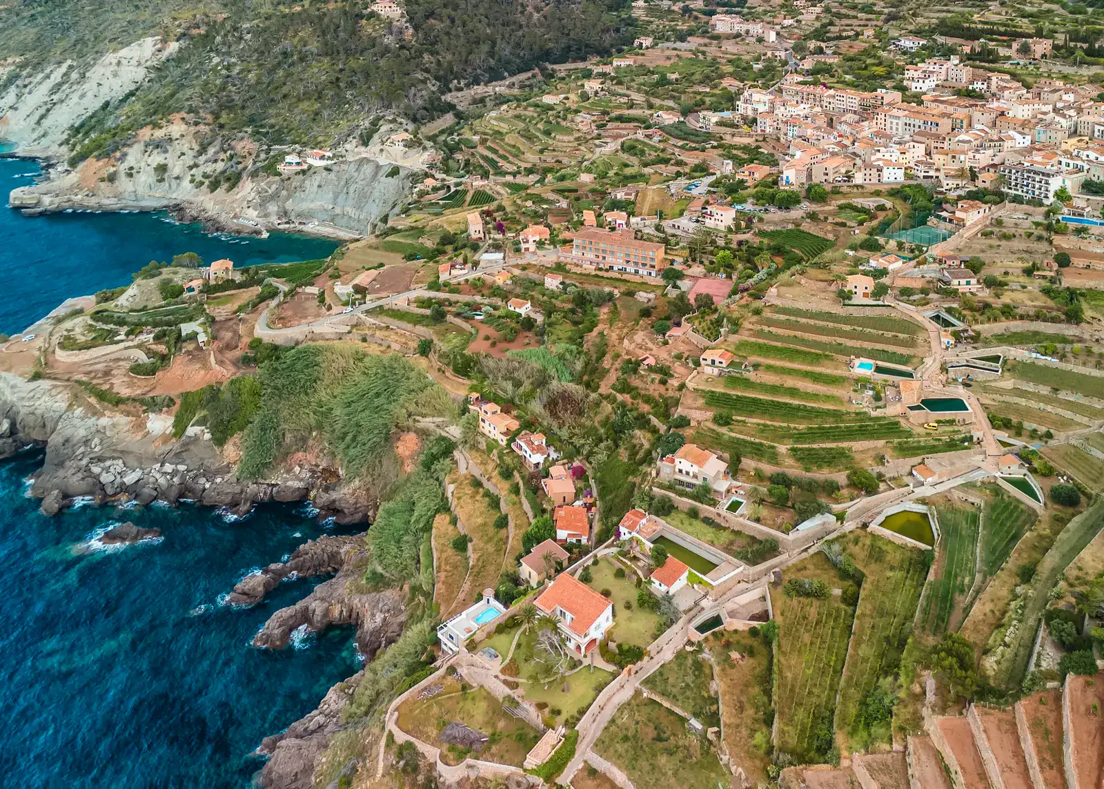 Aerial view of Banyalbufar, a village of beige stone houses with orange roofs built on green terraces on the slope of a cliff by the water, one of the top places to visit in Mallorca.