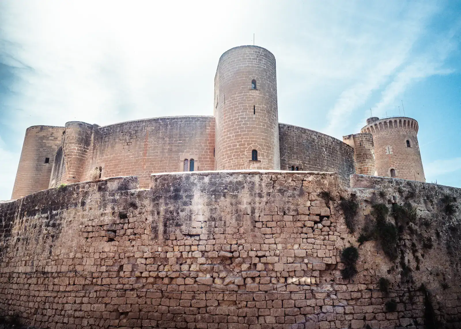 Looking up at the round Bellver Castle from below the outer wall, with the sun shining into the camera, one of the top things to do in Mallorca.