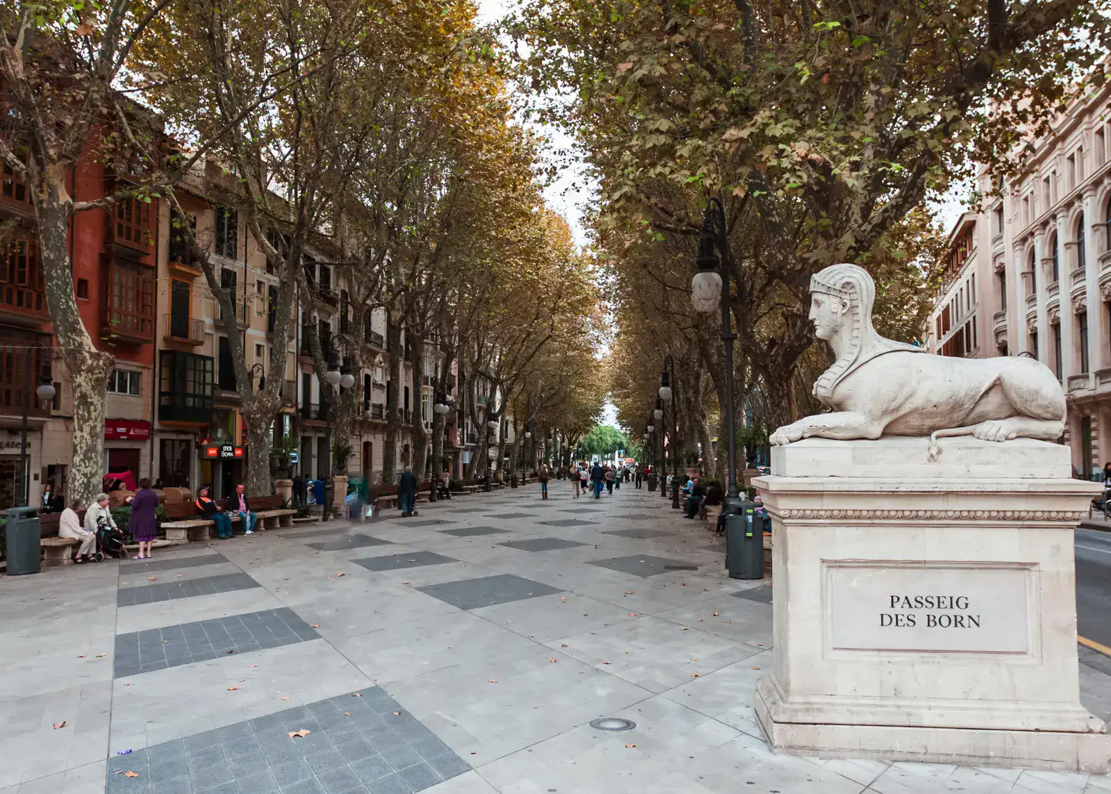 Looking down Passeig des Born, a tiled boulevard lined by trees on both sides, a Egyptian style human/lion state on top of a plinth to the right, people sitting on benches to the left, seen during a one day itinerary of Palma de Mallorca.