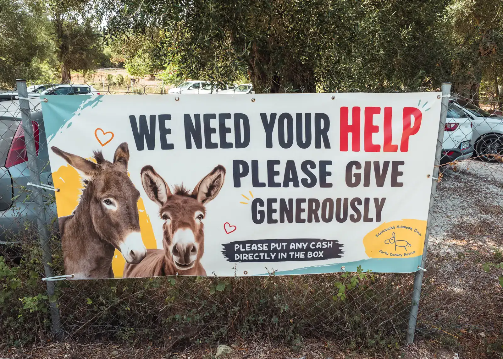 White sign outside the Corfu Donkey Rescue parking, with a photo of two donkey heads and the message: We need your help. Please give generously.