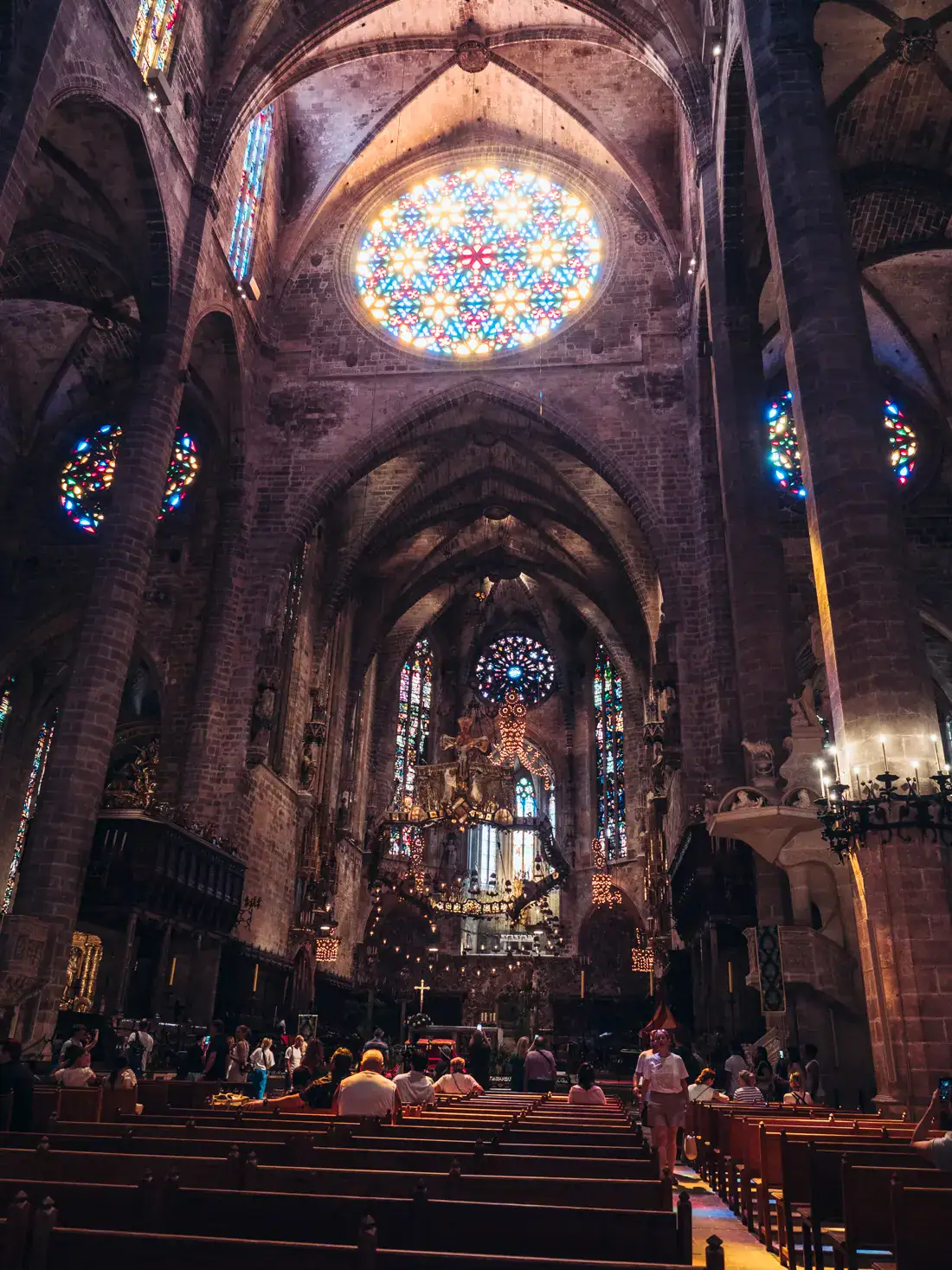 Inside Palma Cathedral, people seated on wooden pews, looking up at a big, round stained glass window, with several arches below, one of the top things to do in Palma de Mallorca.