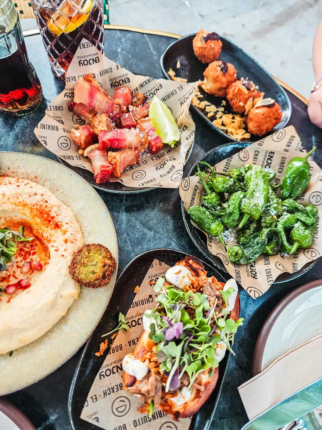 Close up of a table of tapas, round plate with hummus, padr&oacute;n peppers, four round croquetas, pork belly bites and a brioche bun topped with herbs, for dinner during my one day in Palma Mallorca.