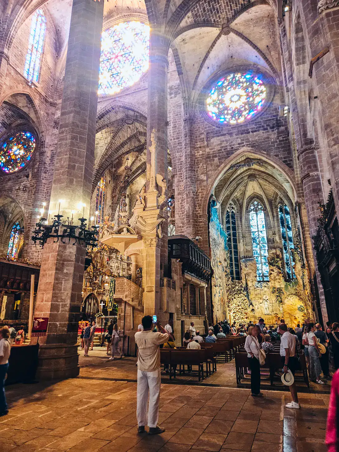 Man taking a photo with his phone inside Palma Cathedral, people standing on the right and seated on wooden pews, looking up at a big, round stained glass window, with several arches below, one of the top things to do in Palma de Mallorca.