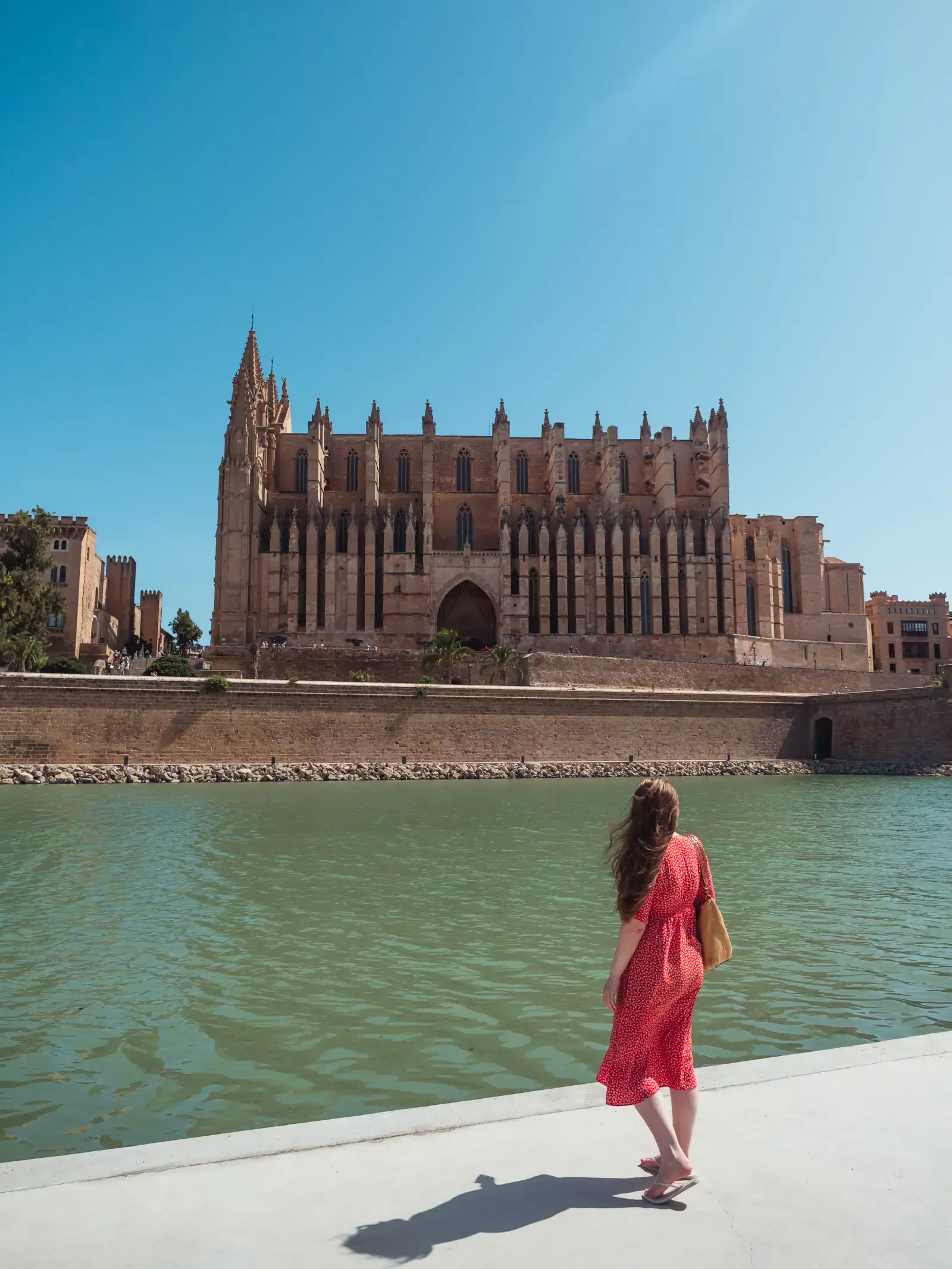 Woman with long brown hair, wearing a red dress, standing on the banks og a green river opposite the majestic Palma Cathedral, a top attraction, built in brown stone on a sunny day in Palma de Mallorca.