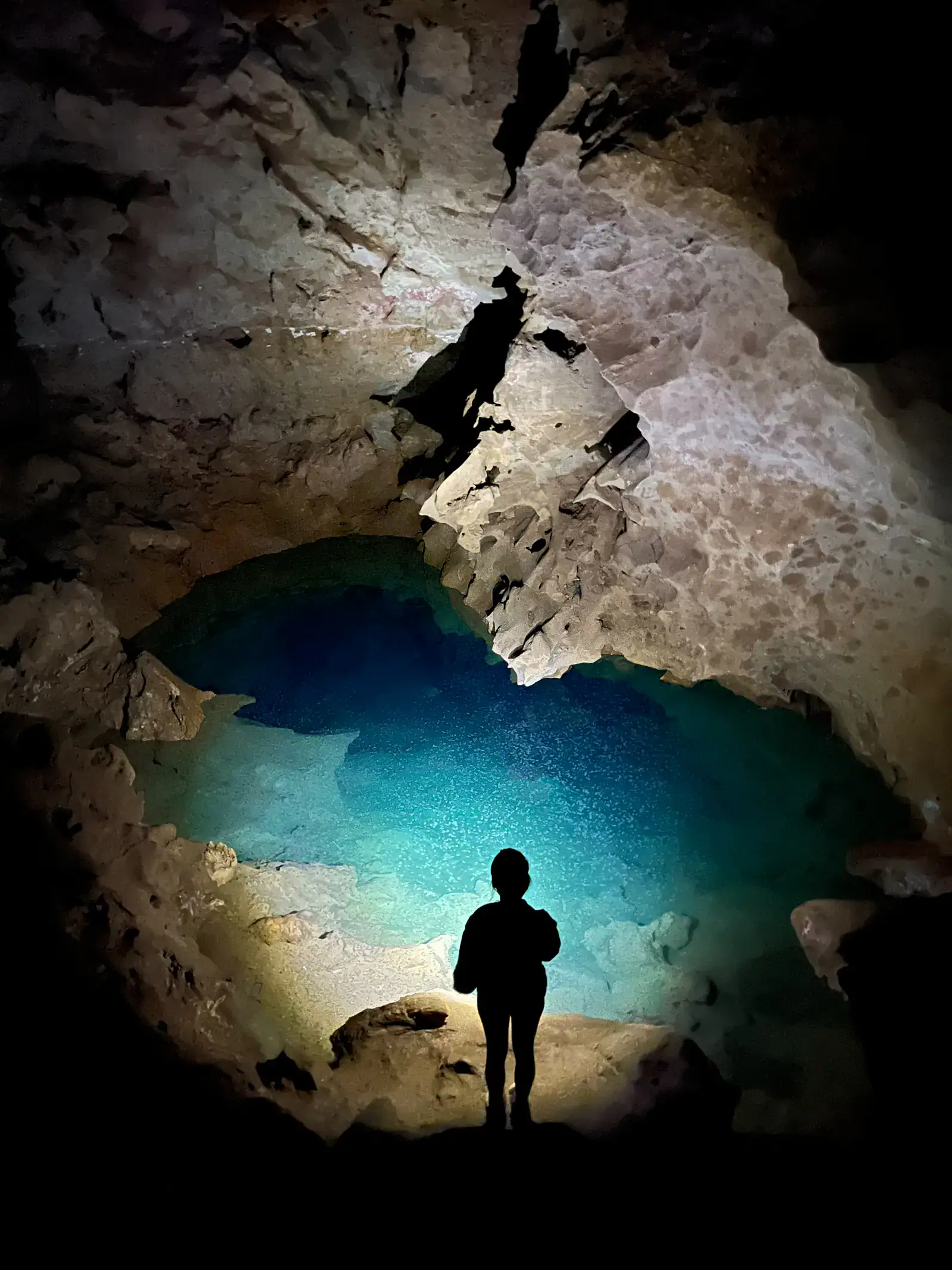 Black silhouette of a woman standing shining a flashlight in a blue pool of water inside Peppersauce Cave, one of the top things to do near Tucson.