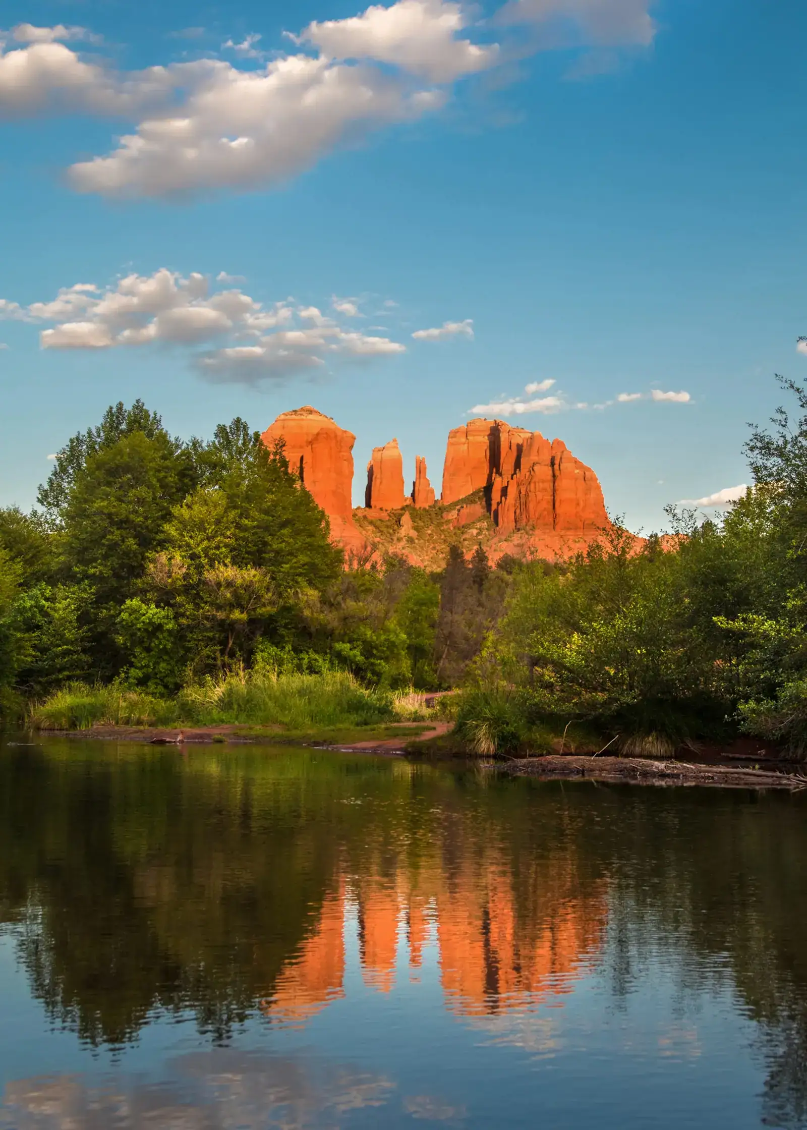 View of Cathedral Rock glowing orange in the sunset, reflecting in waters, surrounded by greenery and a blue sky, why Sedona is one of the best places to visit in the USA in October.