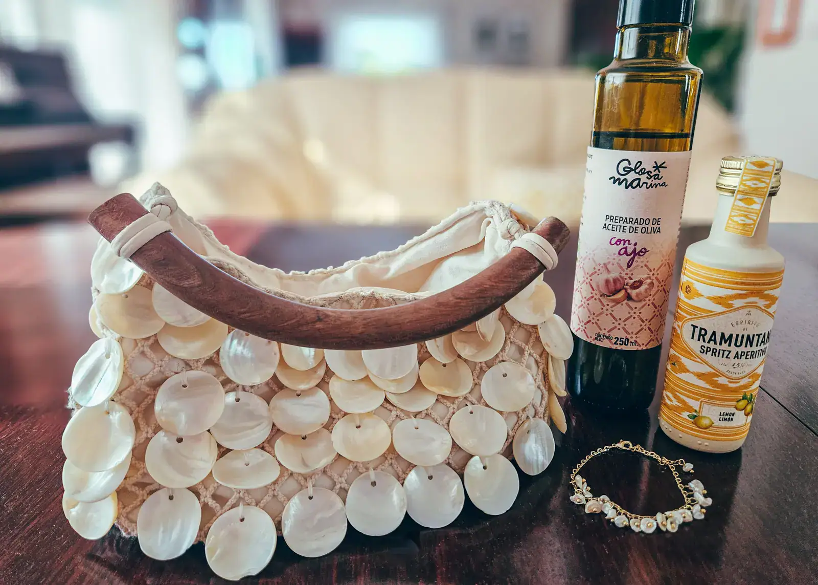 Brown table with a off-white handbag covered in round seashells with a brown wooden handle, gold bracelet with white pearls, dark bottle of garlic olive oil and a small white and yellow liquor bottle, all bought while shopping in Palma.