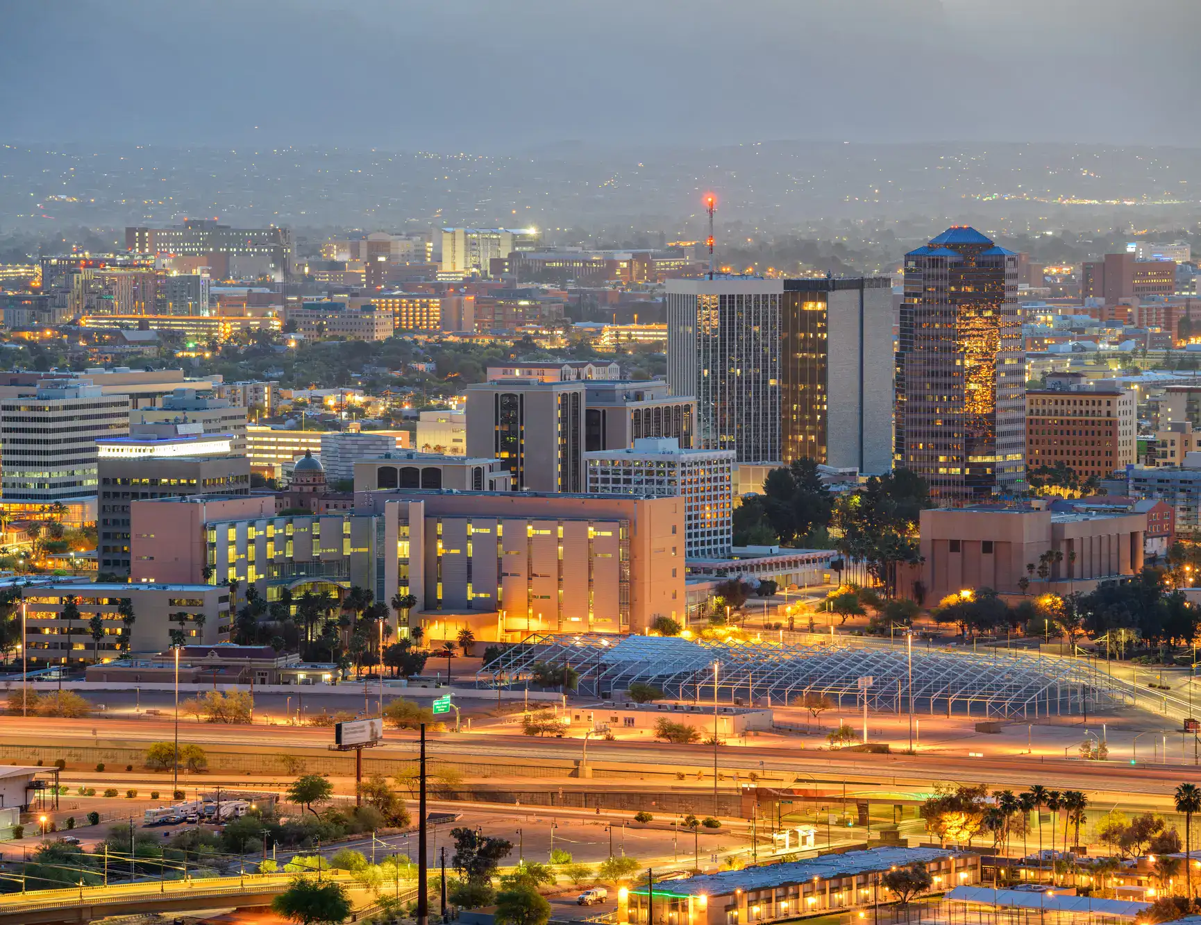 View of Downton Tucson at dawn with orange and yellow light coming from the buildings, train tracks in front, the city stretching out in the background, one of the top things to do in Tucson.