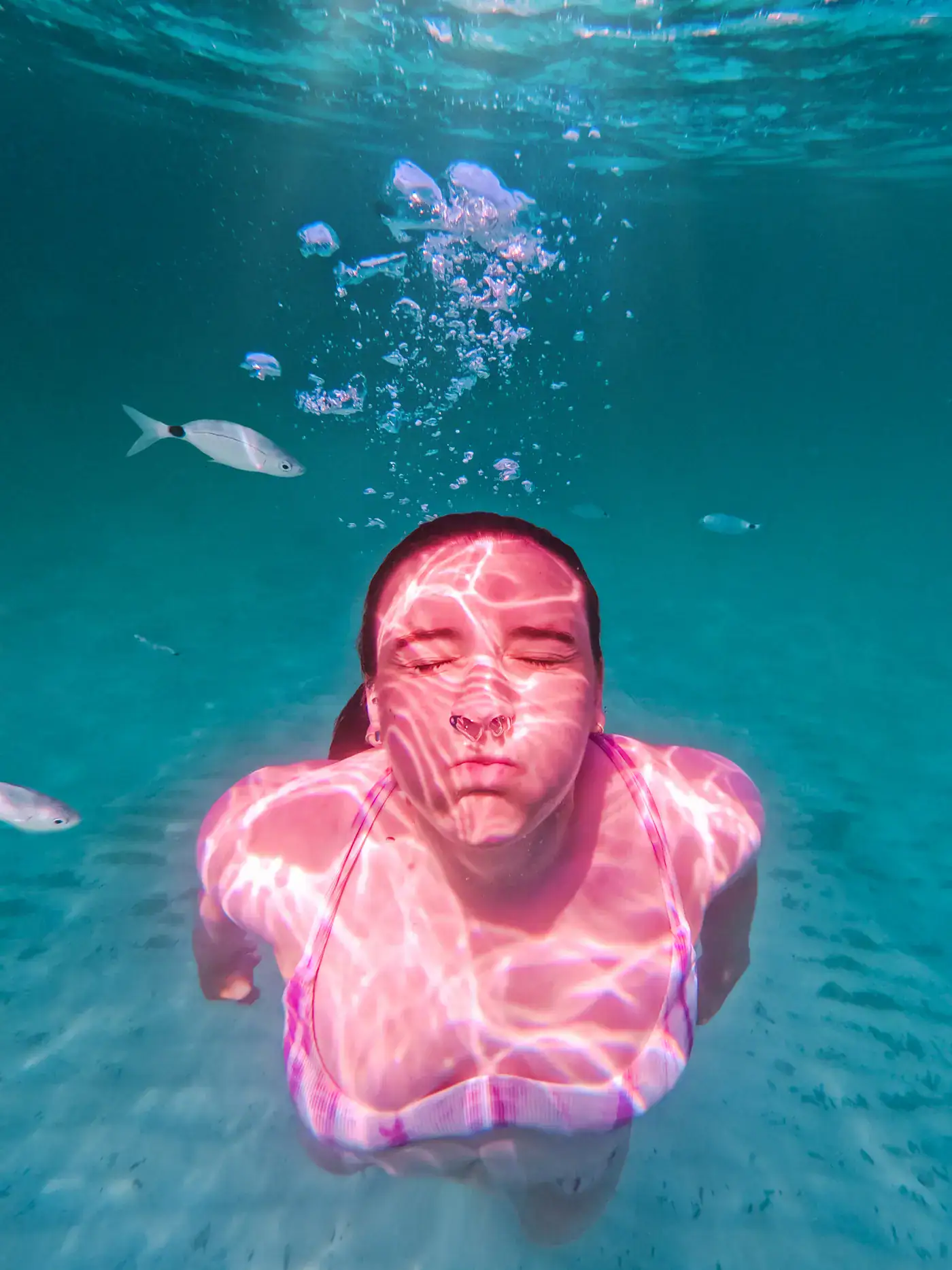 Close up of a woman with dark hair, swimming under water up towards the camera, wearing a white and purple bikini top, blowing out bubbles with her eyes closed, three fishes around her in the azure water at Playa S'Amarador, one of the top things to do in Mallorca.