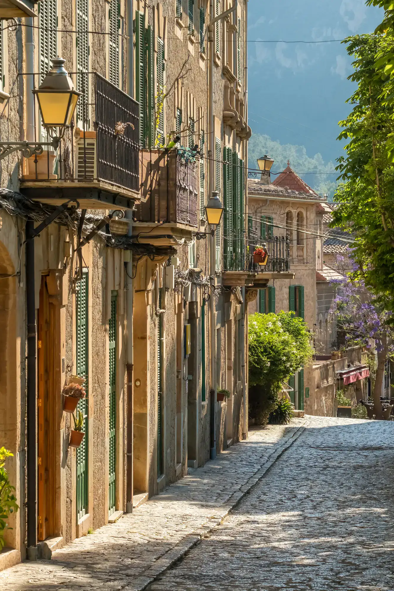 Walking down a cobbled street in Valldemossa, with a grand stone house on the left with green doors and shutters, mountain with greenery in the background, one of the top things to do in Mallorca.