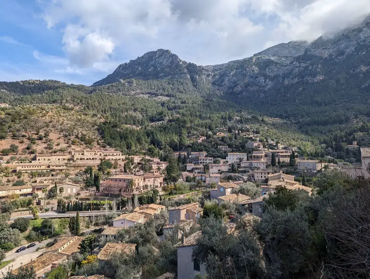 Drone view of Deia, a small village in the mountains made up og beige stone houses surrounded by greenery on a partly cloudy day, one of the best places to visit in Mallorca.