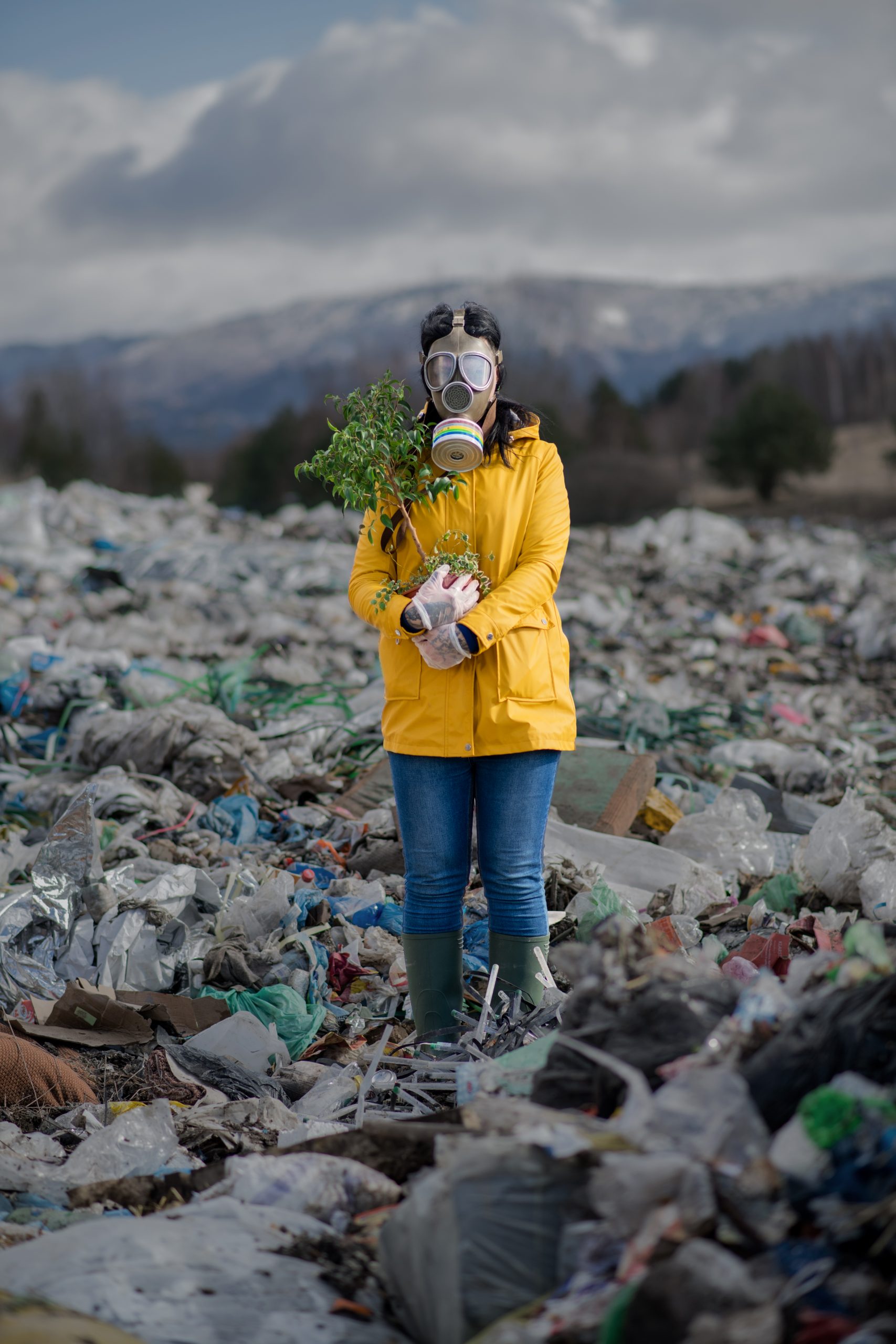 Front view of woman with gas mask holding green plant on landfill, environmental concept.