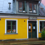 A picture of Hökeriet, an old-fashioned shop and museum in Lund, Sweden. The shop is located in a yellow house on the corner of Sankt Annegatan and Tomegapsgatan, near the open-air museum Kulturen. The shop sells local products such as juice, jam, honey, and candy in paper cones. The shelves are filled with vintage packages from the turn of the 20th century, giving a glimpse of the past. The shop has a red sign above the entrance that says “E.H. Larsson Win- & Diverseaffär”, which was the name of the original owners who opened the shop in 1898. The shop also has a small café where visitors can enjoy coffee and waffles. The street is quiet and cobblestoned.