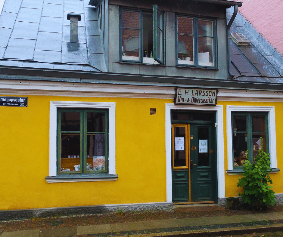 A picture of Hökeriet, an old-fashioned shop and museum in Lund, Sweden. The shop is located in a yellow house on the corner of Sankt Annegatan and Tomegapsgatan, near the open-air museum Kulturen. The shop sells local products such as juice, jam, honey, and candy in paper cones. The shelves are filled with vintage packages from the turn of the 20th century, giving a glimpse of the past. The shop has a red sign above the entrance that says “E.H. Larsson Win- & Diverseaffär”, which was the name of the original owners who opened the shop in 1898. The shop also has a small café where visitors can enjoy coffee and waffles. The street is quiet and cobblestoned.