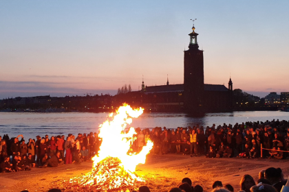 A large bonfire on Riddarholmen, Stockholm, with a view of the City Hall and the water. A large crowd stands around the fire.