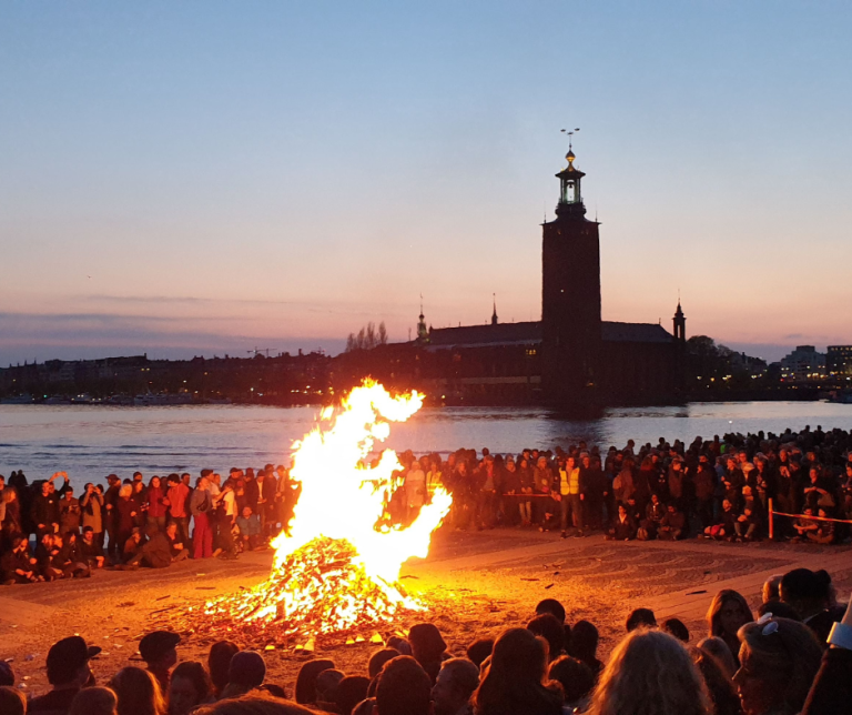A large bonfire on Riddarholmen, Stockholm, with a view of the City Hall and the water. A large crowd stands around the fire.