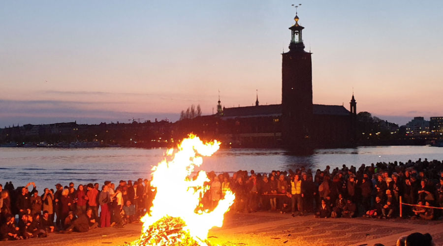 A large bonfire on Riddarholmen, Stockholm, with a view of the City Hall and the water. A large crowd stands around the fire.