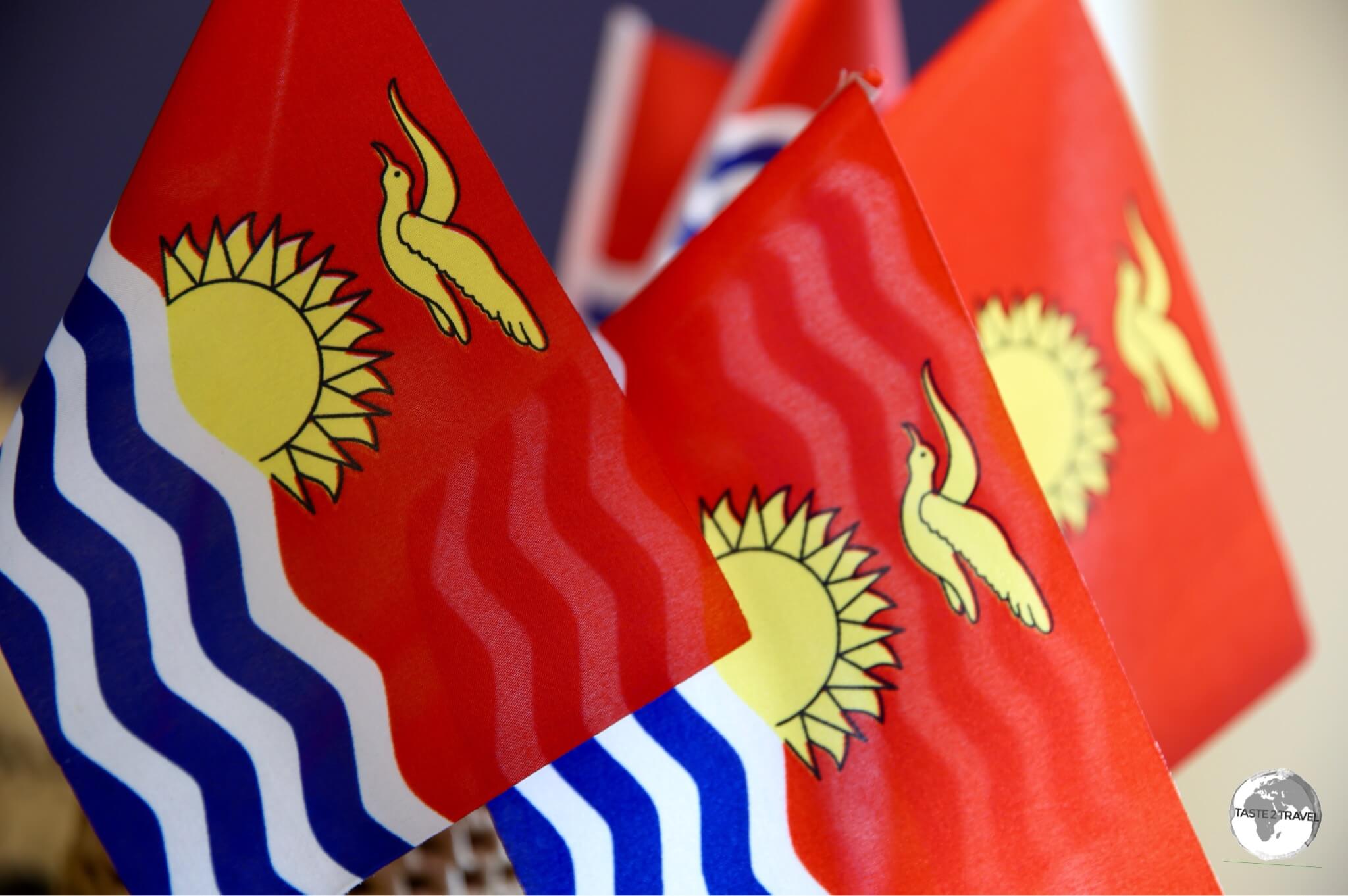 The national flag of Kiribati features a gold Frigate bird flying over a golden sun with three blue/ white bands representing the ocean and the three island groups.