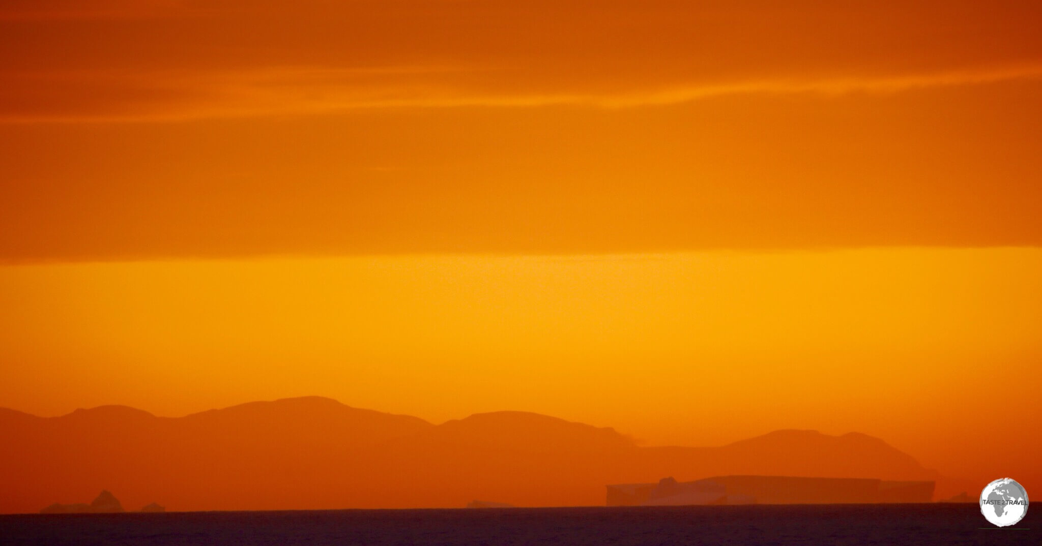 The darkened, rectangular form of a large iceberg stands out against a mountain range in the setting sun.