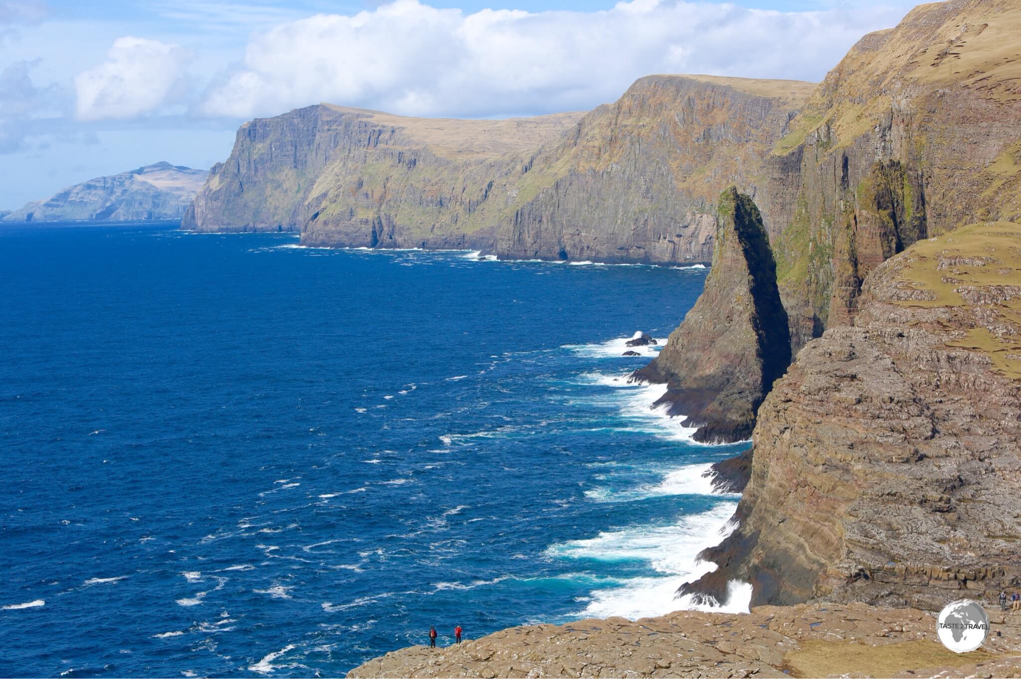 Two hikers are dwarfed by the towering cliffs of the Vágar coast.