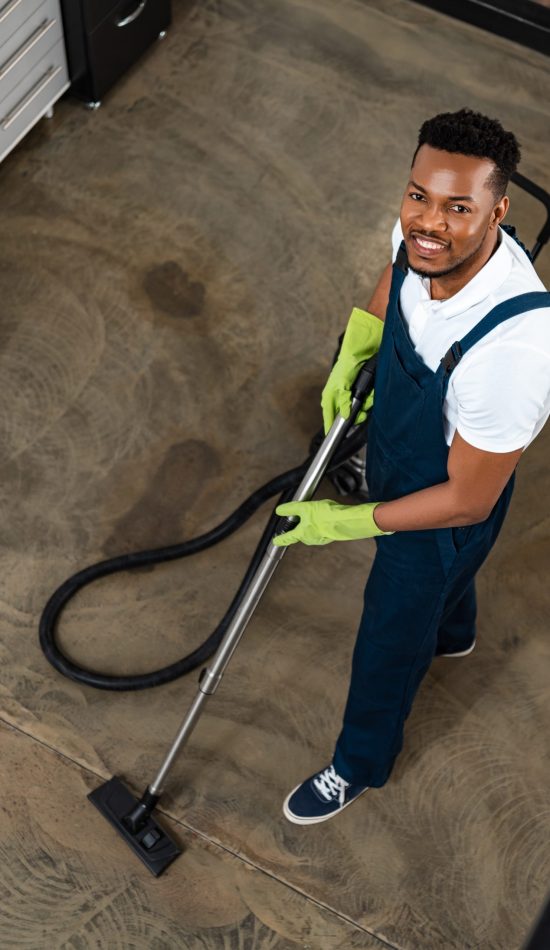 high angle view of smiling african american cleaner vacuuming floor while looking at camera