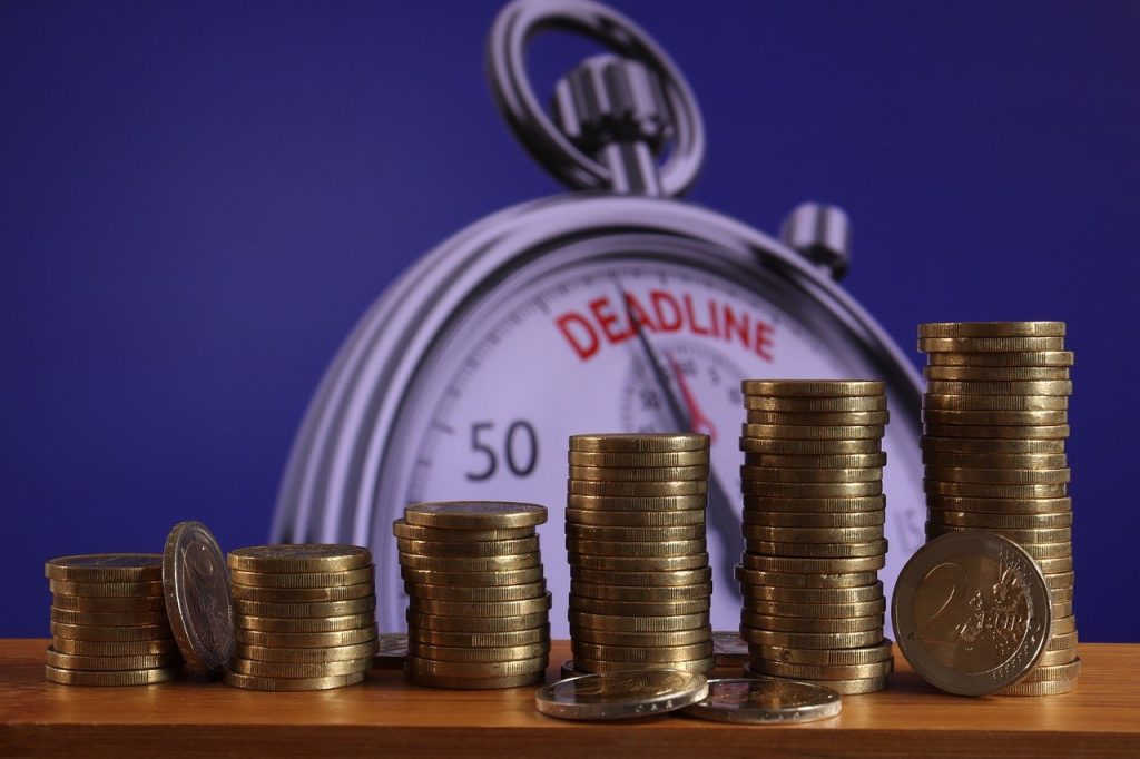 A visual of a pile of coins in front of a table clock signaling a deadline