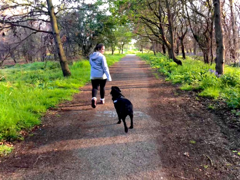 A young woman wearing a jacket and running gear is shown jogging with a dog in a forest