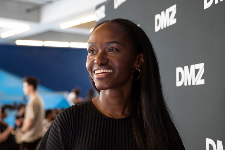 A young Black woman with long black hair, wearing a black sweater smiles standing in front of a banner with the DMZ logo. 