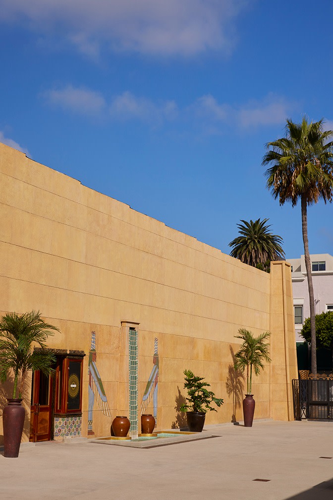 The exterior of a light stone building is decorated with Egyptian-style hieroglyphs; exotic plants in pots are placed along the base of the wall