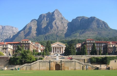 A collection of university buildings, many with red roofs, set against a mountainous backdrop and in front of a grassy field.