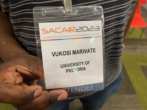 A close-up photo of a person in a striped shirt using their hand to present a conference name tag that says "Vukosi Marivate."