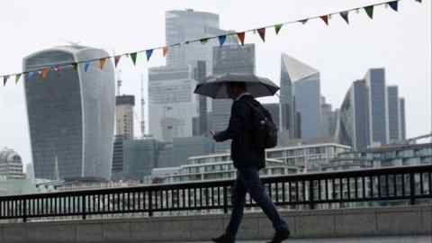 A pedestrian holding an umbrella walks along the Thames river bank in view of the City of London skyline