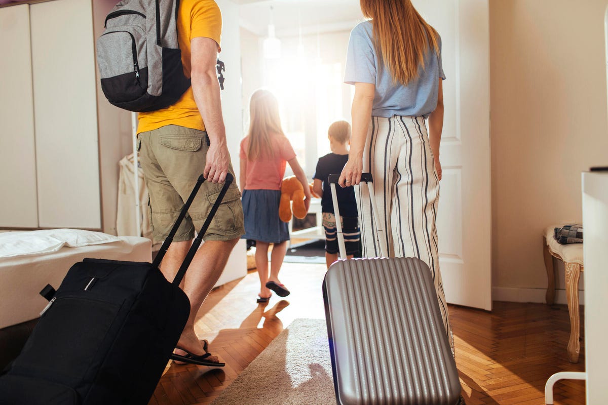 A family with luggage walks through a sunlight front door.