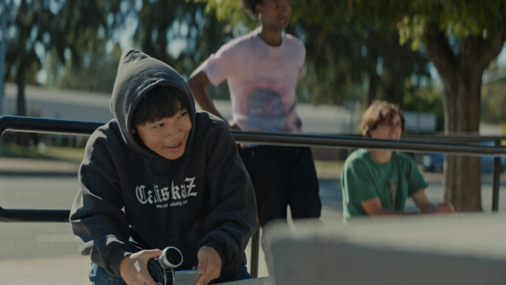 Asian teenage boy outside with friends holding a video camera and smiling.