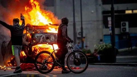 Getty Images Two men covering their face celebrate, one holding a bottle of alcohol, while staring at the flaming wreckage of what was once a police car, now a fireball