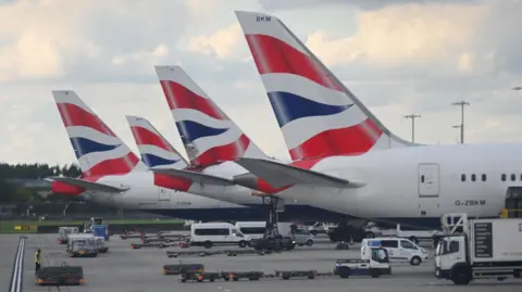 The tailfins of four British Airways planes on the tarmac at Heathrow Airport. The planes are white with a stylised part of the union jack on their tailfins