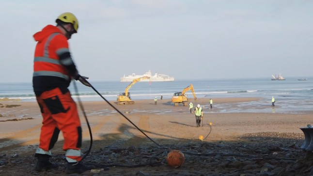 Google's Grace Hopper fiber cable being laid on a beach by a man in orange work gear, with workers in the background