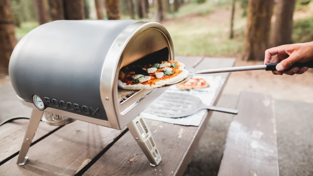 En pizza fjernes fra en Roccbox Roccoboxpizzaovn på et picnicbord i en skov, personens hånd holder en pizzaspade.